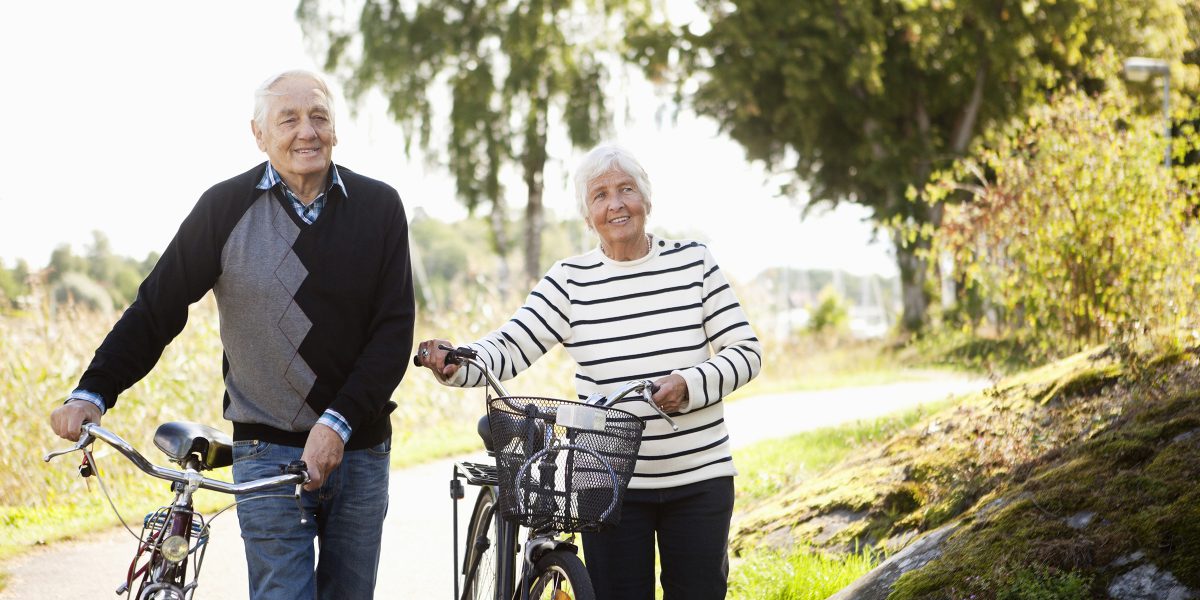 Ein eldre mann og ei eldre kvinne gåande med kvar sin sykkel