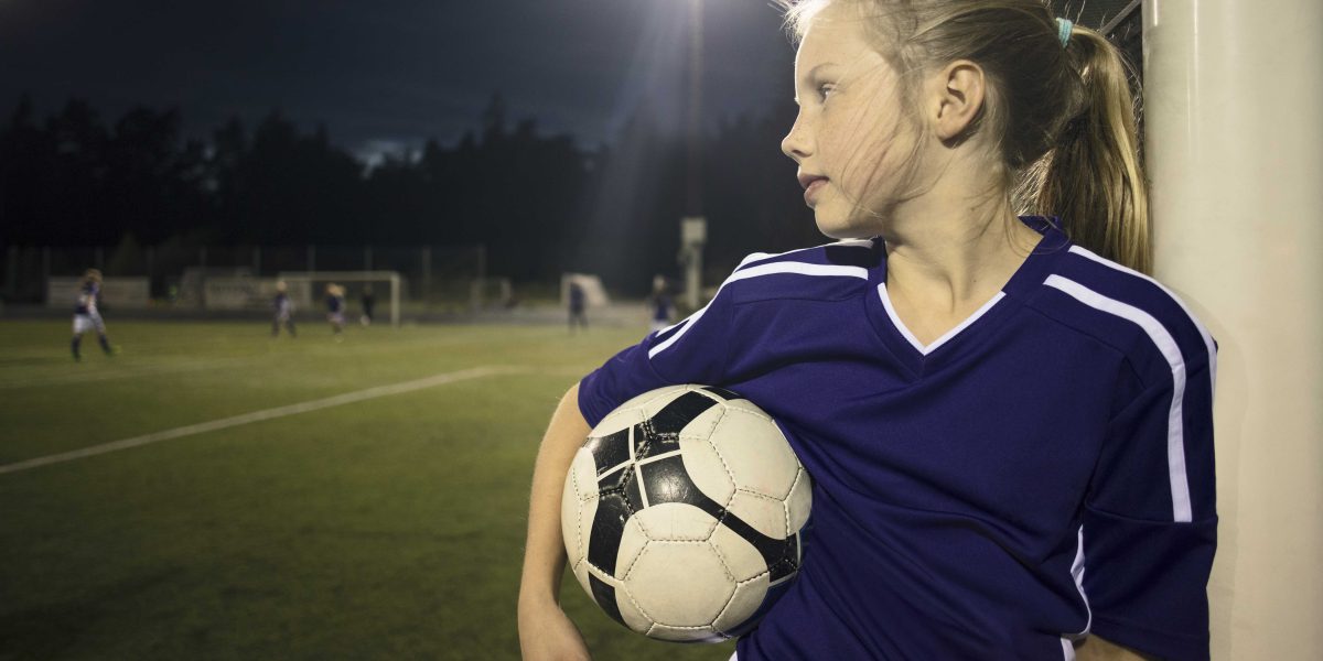 Jente med fotball ser på andre ungdommer som spiller fotball ute på banen.